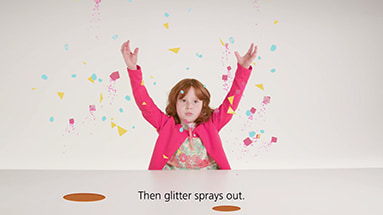 Still of the ubs film showing a girl with red hair and a pink cardigan sitting on a white table in a white space with colorful geometrical shapes and glitter flying around her with a subtitle saying "Then glitter sprays out.".