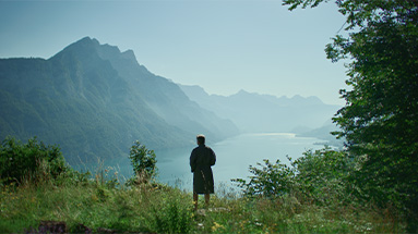 Still of the hüsler nest film showing a man standing in his garden and looking at the lake and the mountains in the background.